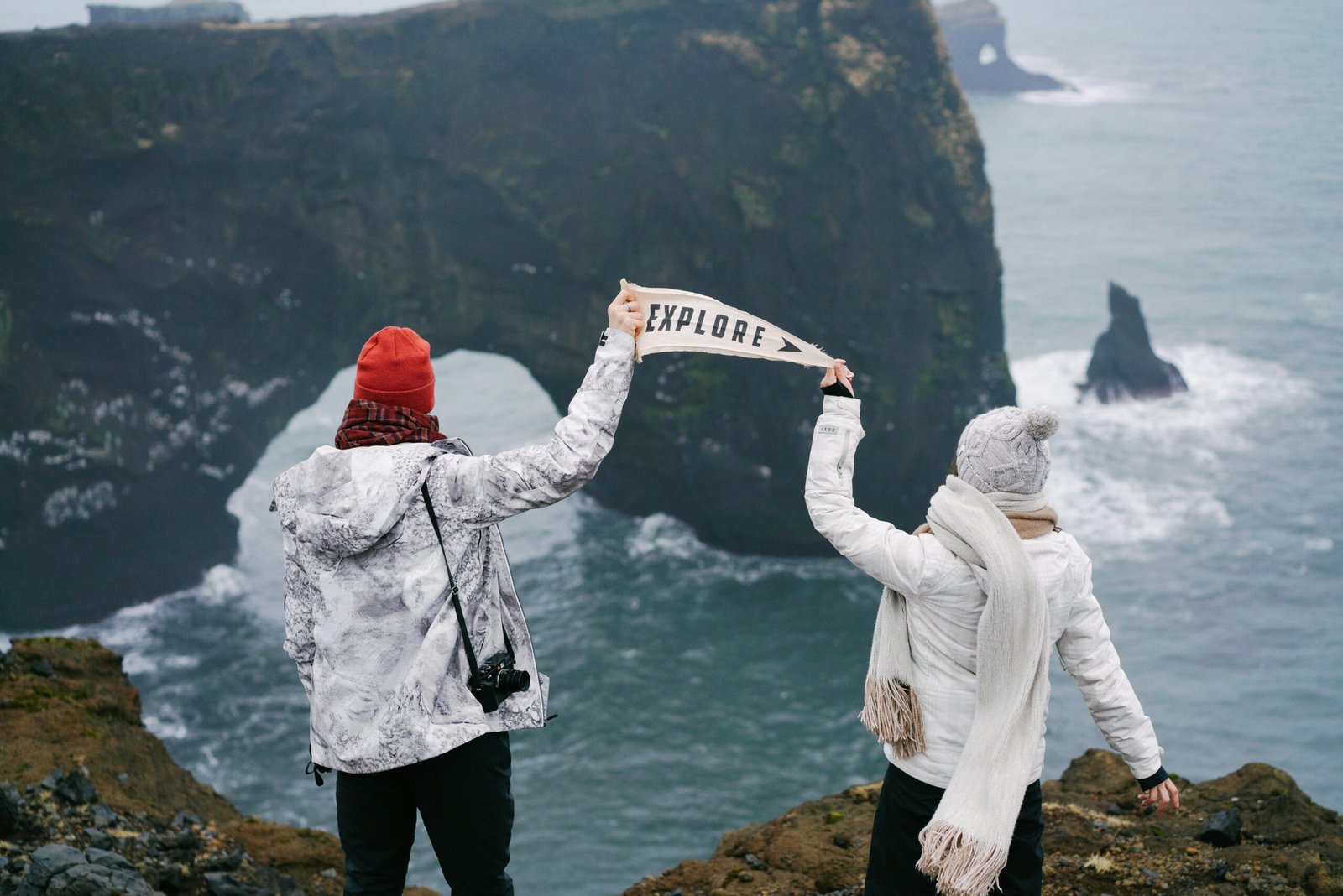 Two hikers stand on a cliff edge holding an 'Explore' sign, overlooking a dramatic coastal arch.