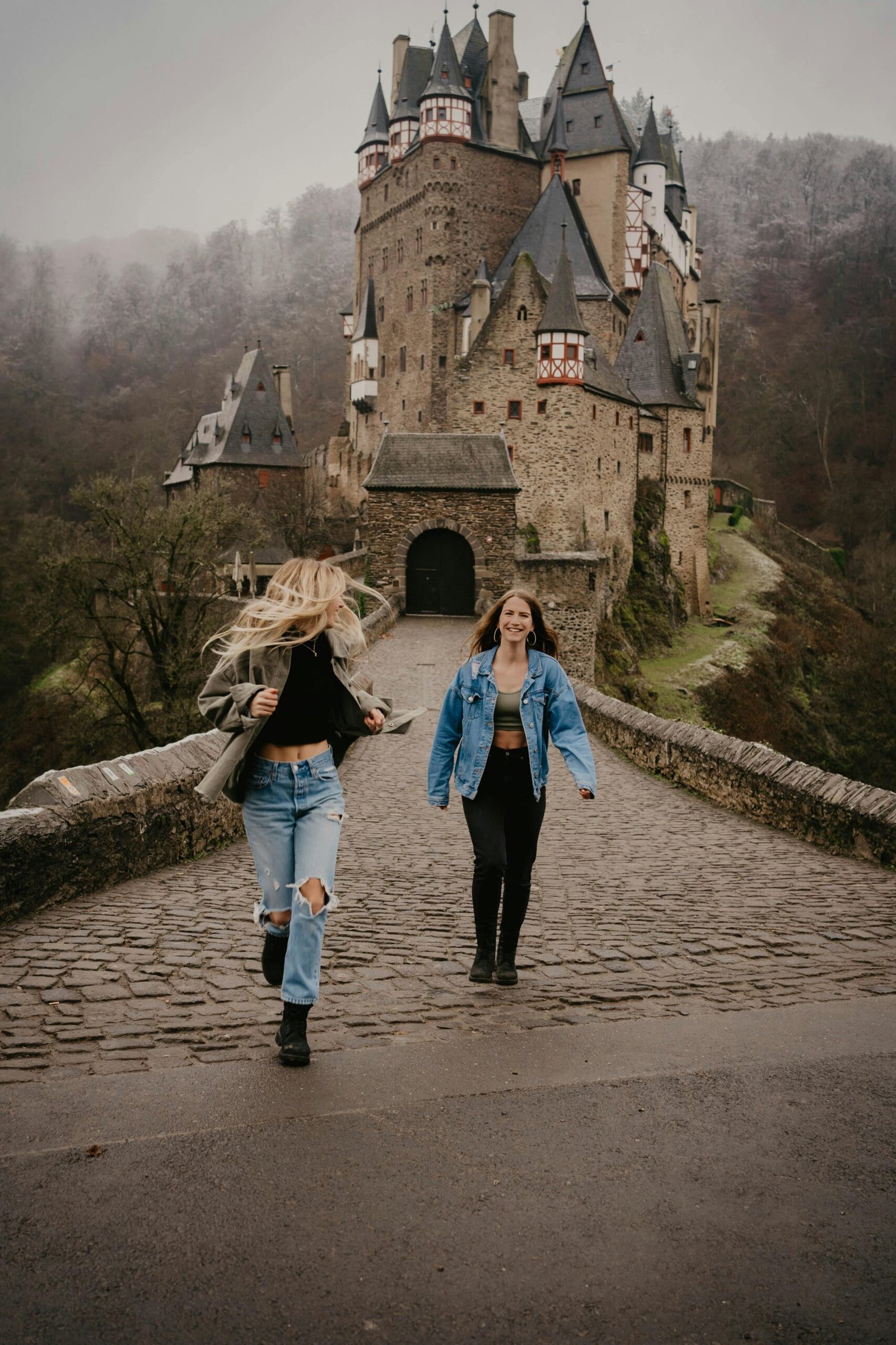 Two women walking on cobblestone bridge towards Eltz Castle surrounded by fog